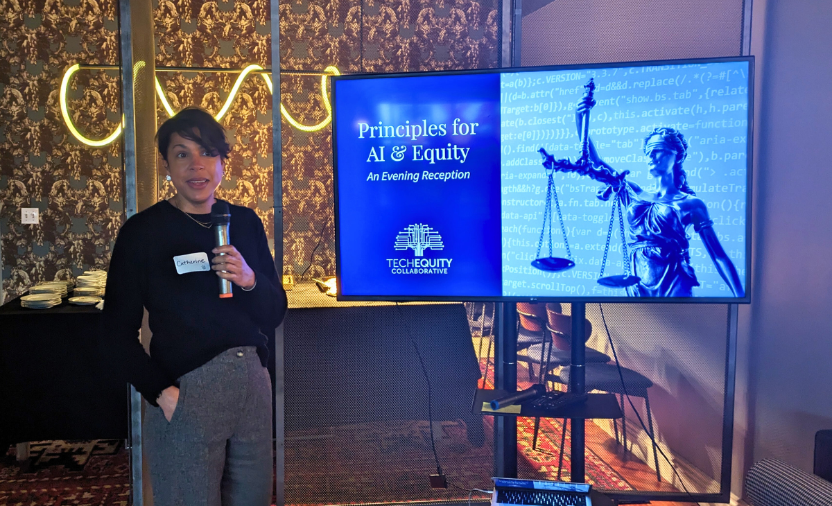 TechEquity Founder and CEO Catherine Bracy standing with a microphone in hand and there's a TV with a presentation just behind her that says, Principles for AI and Equity: An Evening Reception.